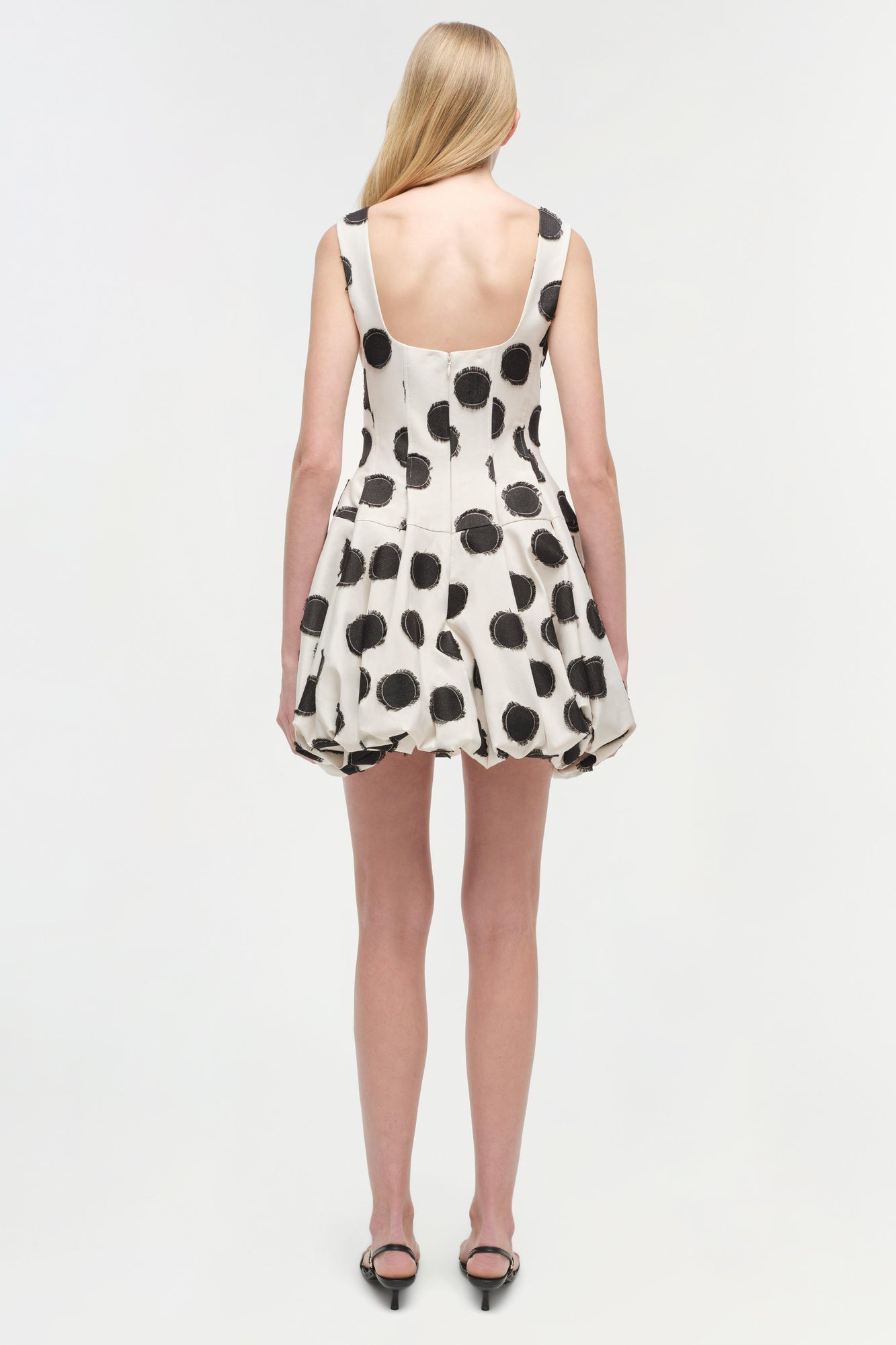 Sleeveless mini dress with black polka dots on cream fabric, open back design, shown from behind on model.