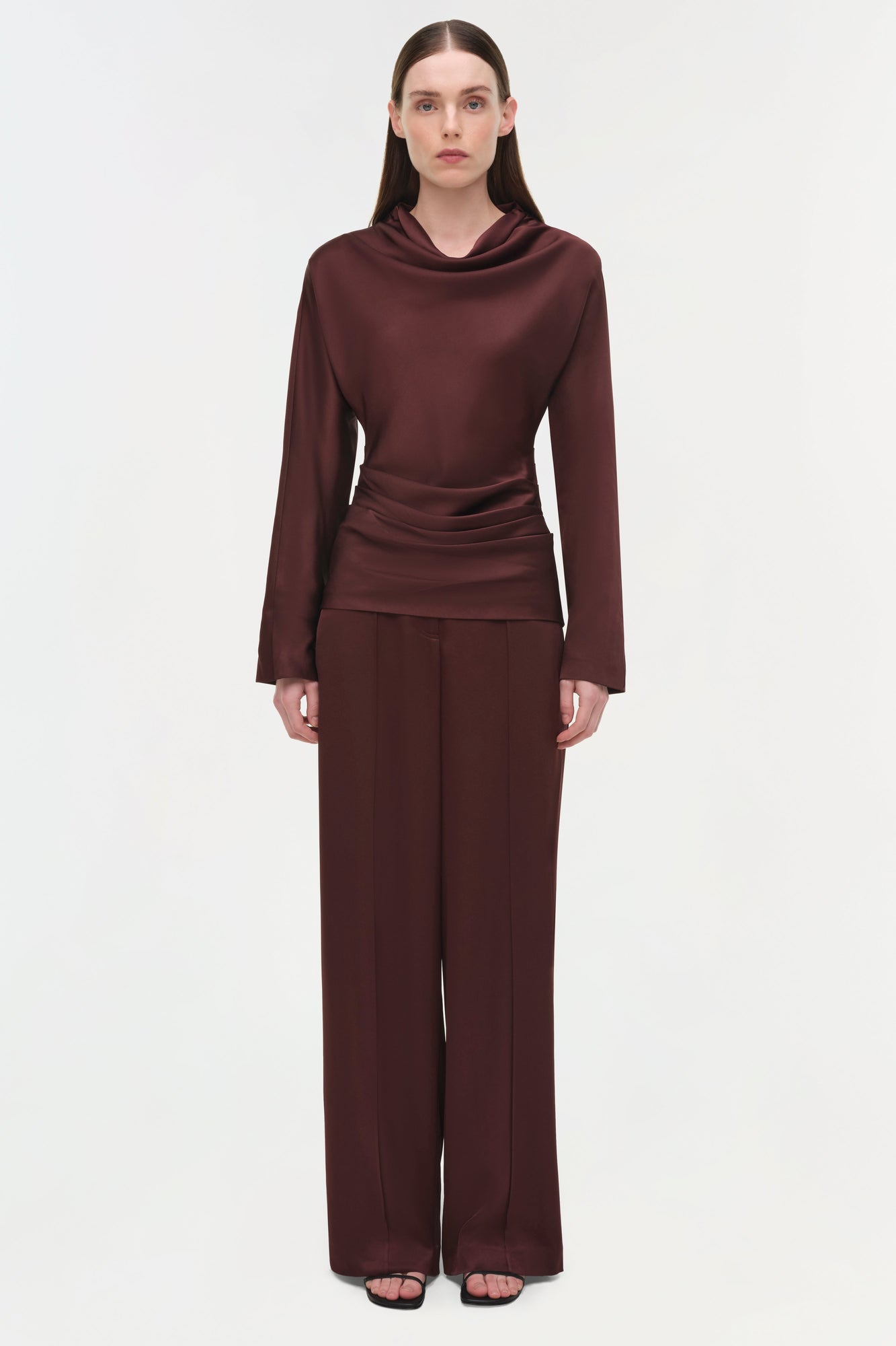 Woman wearing burgundy satin draped long-sleeve top with cowl neckline and ruched waist detail paired with wide-leg pants.
