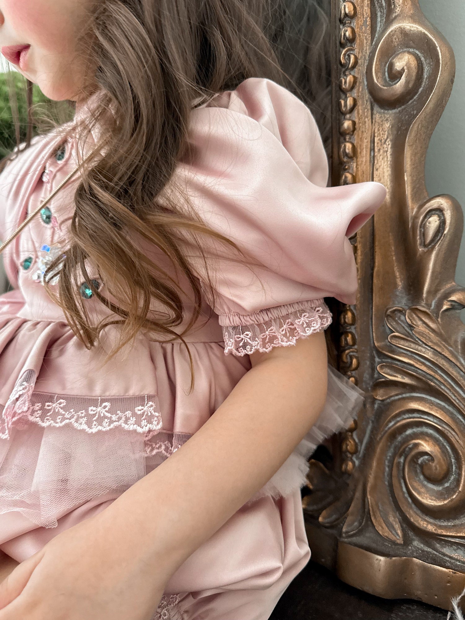 Close up of pink satin romper with ruffled bodice, lace trim, and gem embellishments