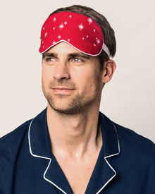 A man in navy pajamas with white trim and a red sleep mask featuring white stars looks aside against a simple backdrop, displaying Petite Plume’s Adults Flannel Sleep Mask in Starry Night, a perfect gift for any occasion.