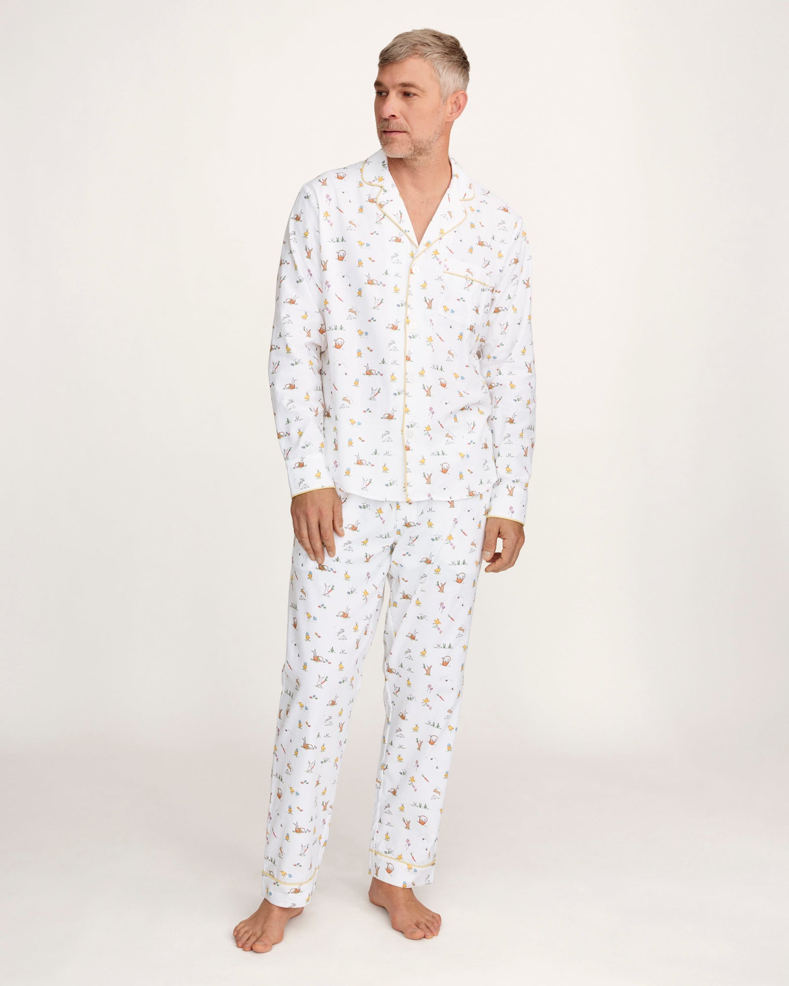 A man with short gray hair wears the Men's Twill Pajama Set in Easter Frolic—white pants and a long-sleeve top with a small, colorful print—standing barefoot against a plain light background.