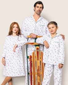 A man and two kids pose with a wooden sled, all wearing Petite Plumes Mens Flannel Pajama Set in Apres Ski. The girls red hair contrasts with their festive 100% cotton pajamas, while the boy shares brown hair similar to the mans short dark hair and facial hair.