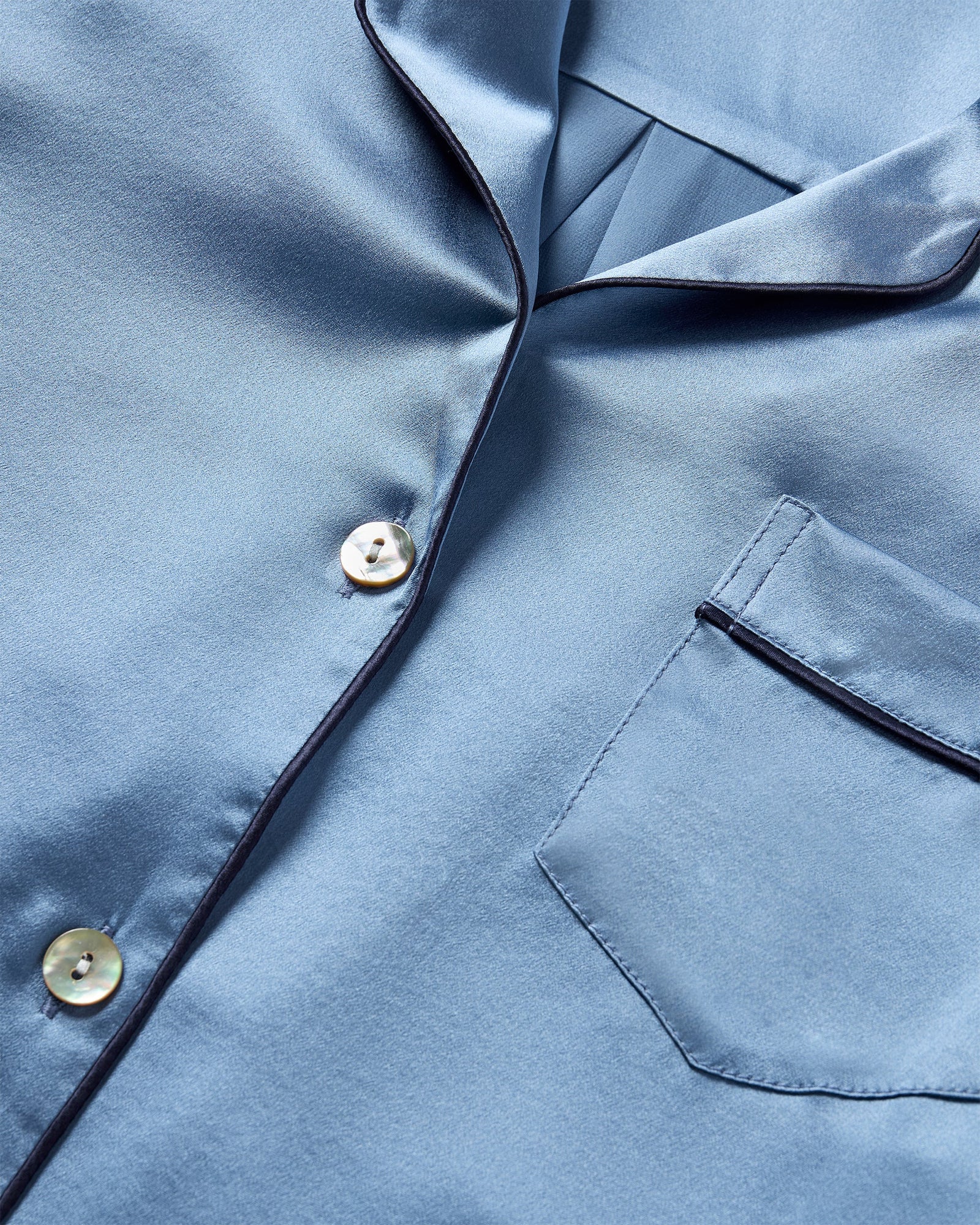 The Men's Silk Pajama Set in Mystique Blue features a button-up long-sleeve top with a chest pocket and matching elastic-waist pants, both trimmed with dark piping.