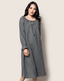 Against a plain background, a woman poses in the Womens Flannel Delphine Nightgown by Petite Plume. Its gray with long sleeves, made of 100% cotton soft flannel, and features a drawstring neckline and loose fit. She stands with one hand on her hip, smiling softly.