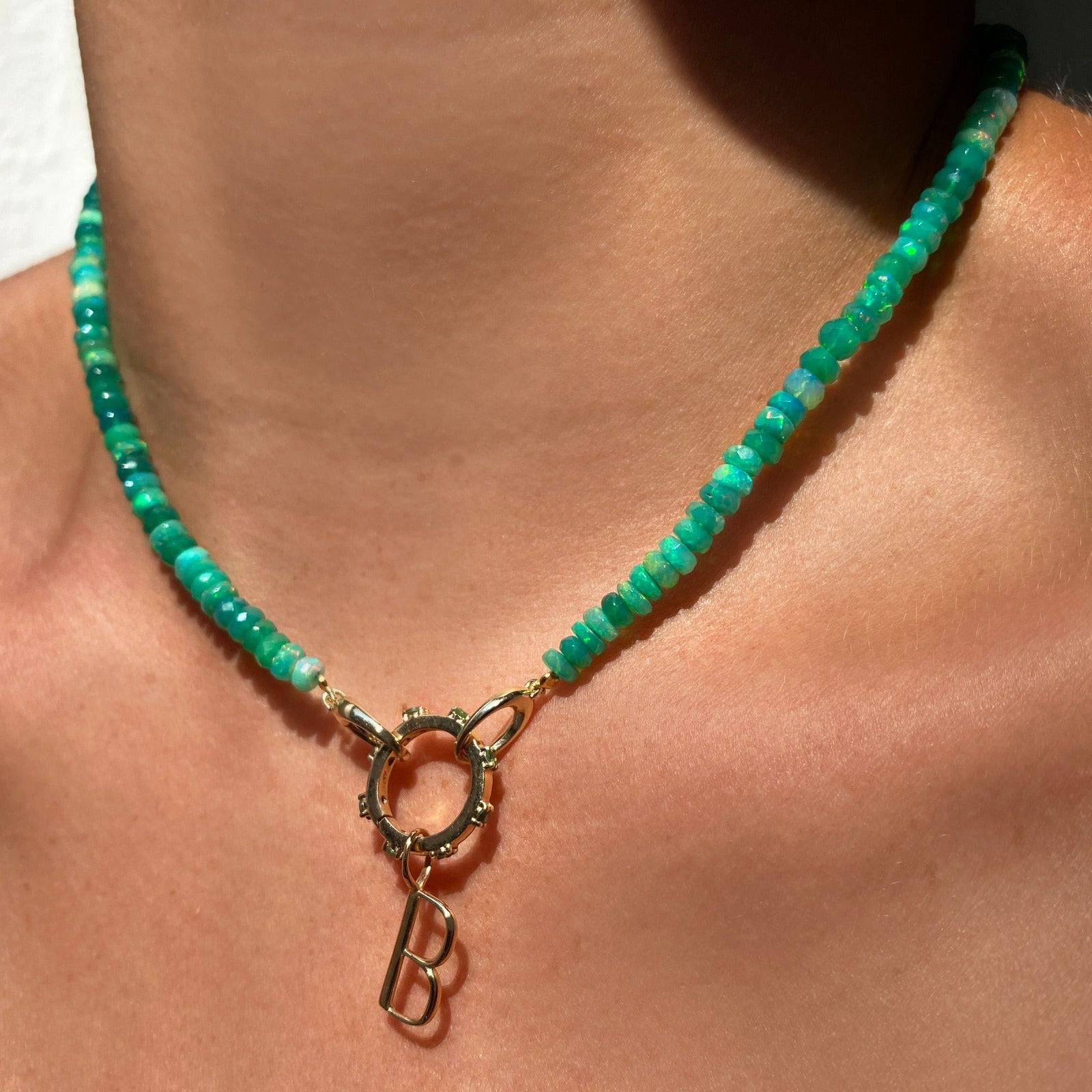 Shimmering beaded necklace made of faceted opals in shades of green on a gold linking ovals clasp. Styled on a neck layered with a round charm lock and B letter charm.