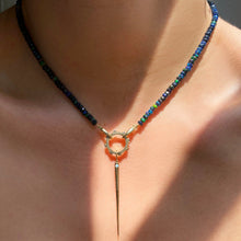Shimmering beaded necklace made of faceted opals in shades of black, green, and blue on a gold linking ovals clasp. Styled on a neck layered with a rounded charm lock and quill spike charm.