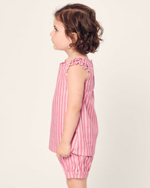 A young child with curly brown hair stands in profile, facing left, wearing the Girls Twill Amelie Short Set in Love Lines by Petite Plume. The set features a pink and white striped sleeveless top with ruffled edges and matching cotton shorts. The background is plain and neutral.