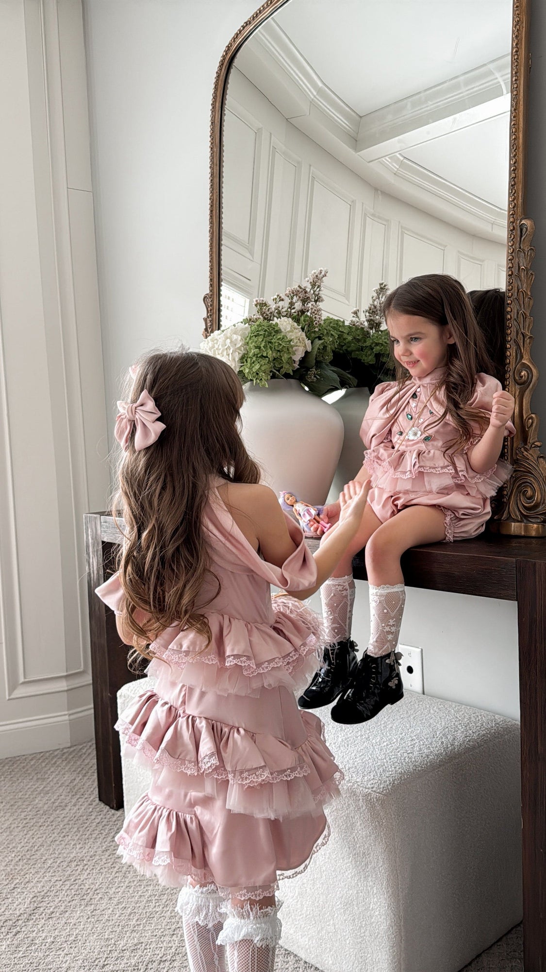 Two girls wearing blush pink satin rompers with ruffled bodice, lace trim, and gem embellishments