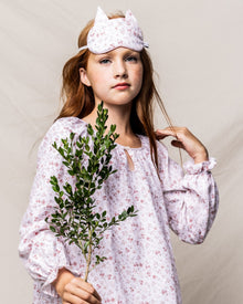 A young red-haired girl wears Petite Plumes Kids Kitty Sleep Mask in Dorset Floral. Holding green leaves against a plain backdrop, she embodies the ideal gift for fans of whimsical bedtime accessories.