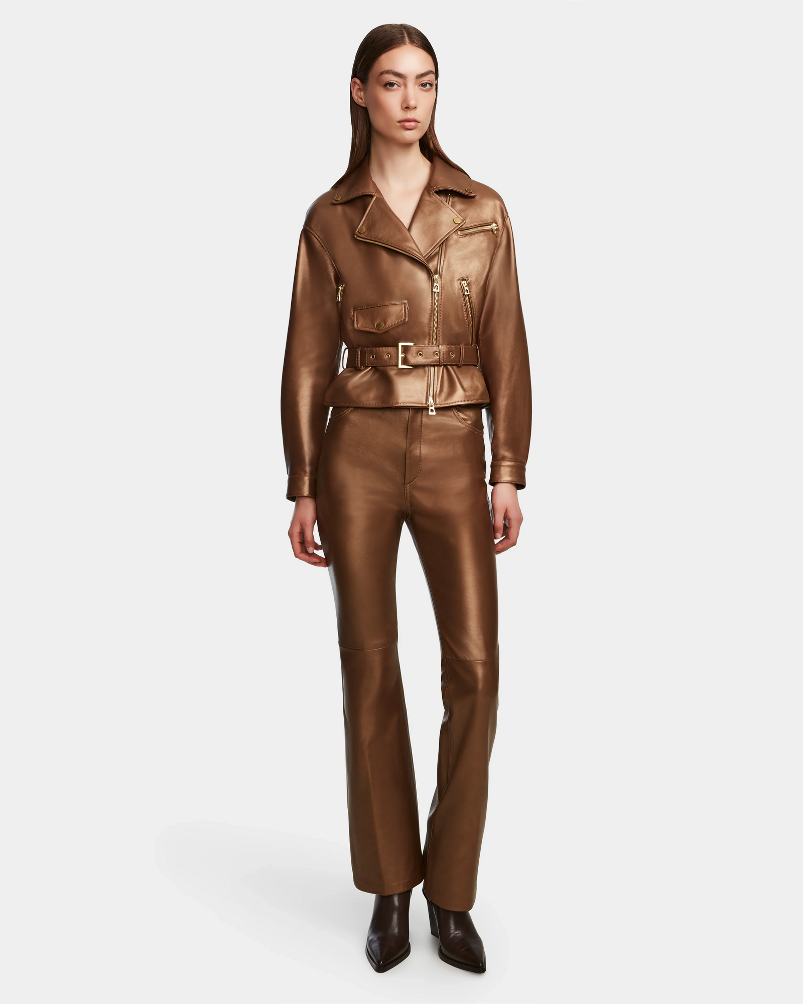 A woman wears the Rudsak ARIELLE - 8124052 Antique Bronze metallic brown relaxed-fit jacket with a belted waist and matching high-waisted leather pants, paired with pointed-toe boots. She has straight hair and a neutral expression against a plain background.
