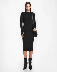 A woman faces forward wearing the Rudsak AGNES - 8524551 fitted long-sleeve black stretch dress, paired with black ankle boots and a black shoulder bag. She has straight brown hair and poses against a plain white background.