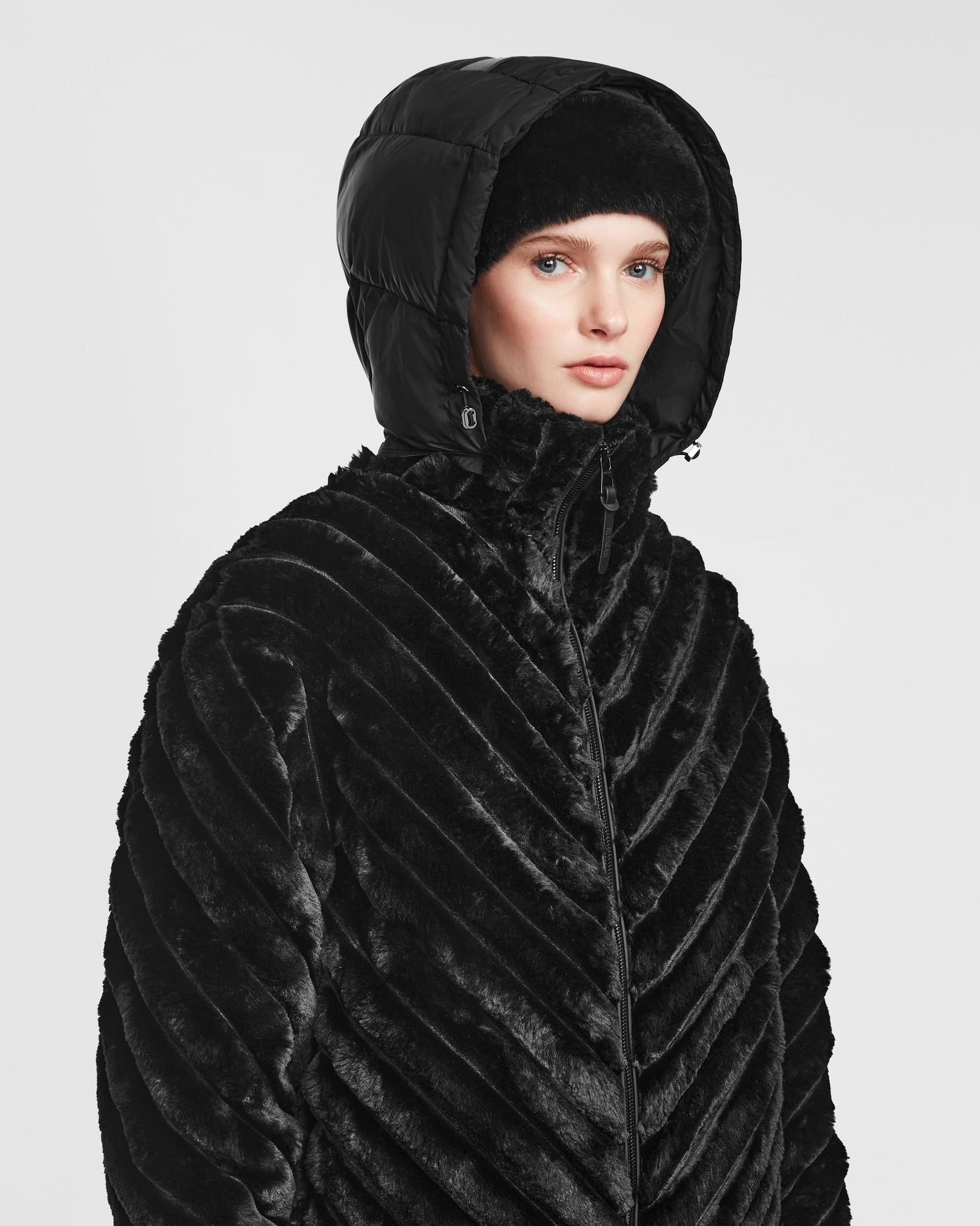 A person models the Rudsak ANASTASIA - 8124804 Black coat, featuring a textured, fur-like finish with diagonal stripes and a puffy hood, against a plain white background.