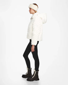 A person in profile wears the Rudsak ANASTASIA - 8124804 Iceland white velvet puffer jacket with a matching faux fur headband, black leggings, and tall dark lace-up boots with cream accents, set against a plain white background.
