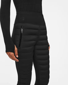 A person models the Rudsak ARIL M - 8522514 BLACK womens quilted pants with zipper pocket and fitted long-sleeve black top, embodying après-ski chic, shown from mid-torso to upper thighs against a plain white background.