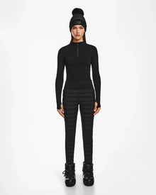 A woman faces forward against a white background, wearing the Rudsak ARIL M - 8522514 BLACK: a chic black fitted long-sleeve top paired with quilted pants, a pom-pom beanie, and winter boots for an après-ski look.