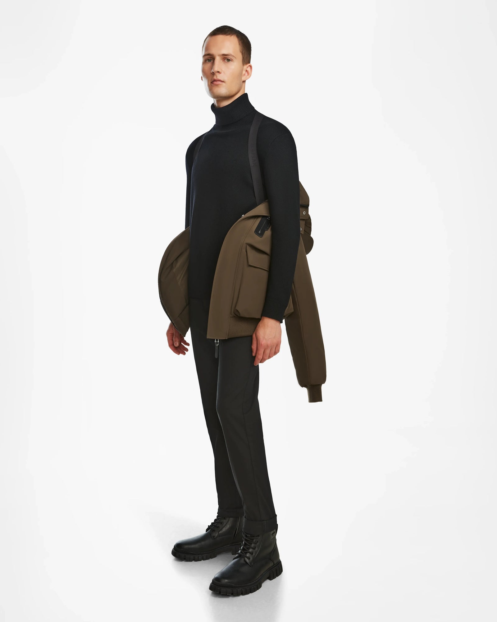 A man poses against a white background in urban style, dressed in black and holding the Rudsak ASH - 6124599 Army mens down bomber jacket draped over his left arm, with a neutral expression.