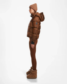A person stands in profile against a white background, wearing the Rudsak ASHLYN S - 8124565 Antique Bronze womens down puffer jacket with hood, paired with matching leggings, a knit beanie, and ankle-height winter boots.