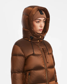 A person wearing the Rudsak ASHLYN S - 8124565 Antique Bronze womens down puffer jacket with the hood up and a beige knit beanie, looking to the side against a plain white background.
