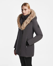 A blonde woman wears the Rudsak AURORE - L123561 Grey fitted winter coat with a large faux fur-lined hood, standing against a white background. She looks at the camera with a neutral expression, hands in the coat pockets.