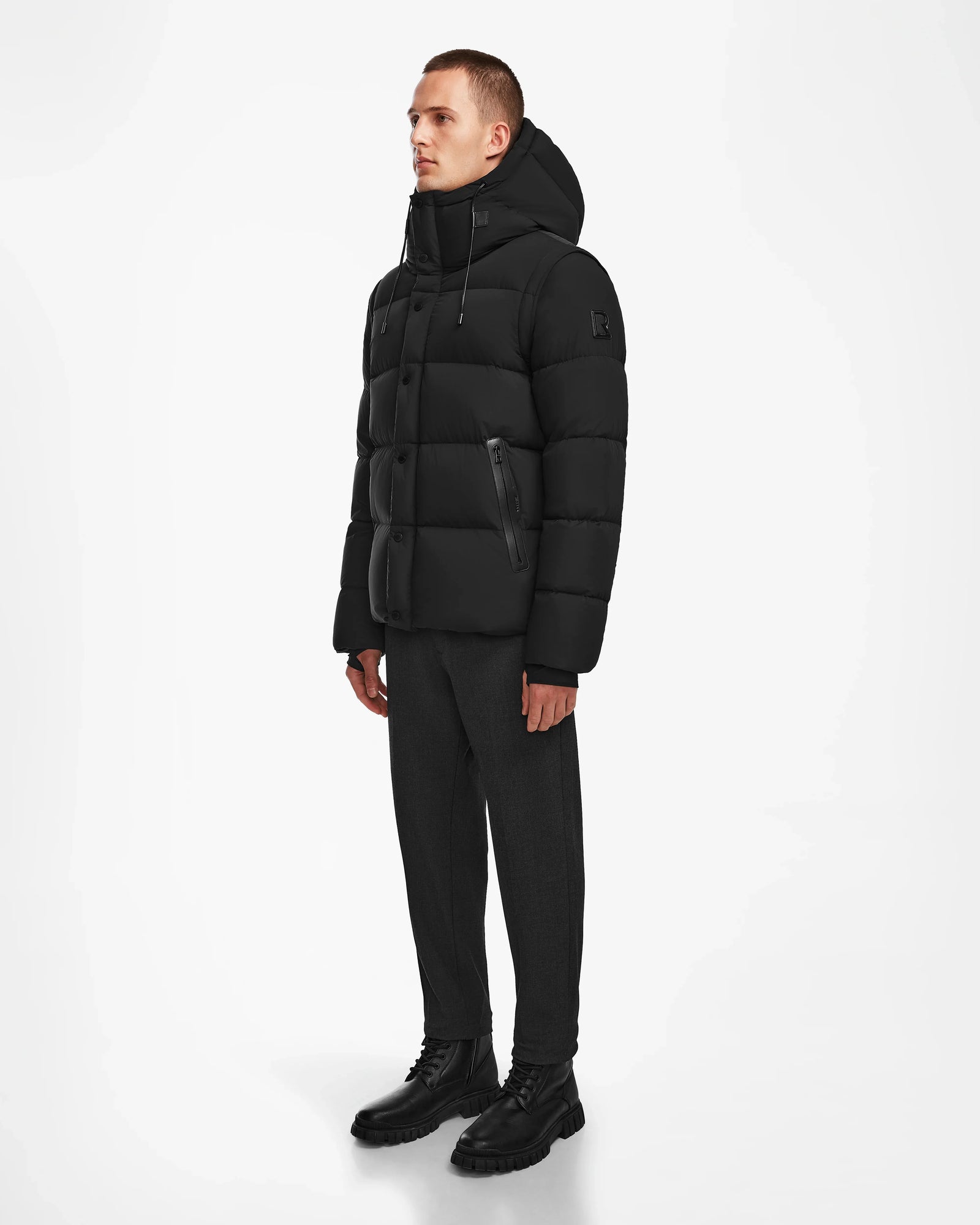 A person stands against a white background wearing the Rudsak BOBBY - 6124506 Black men’s down puffer jacket, dark pants, and black boots, looking slightly to the side with arms relaxed at their sides.