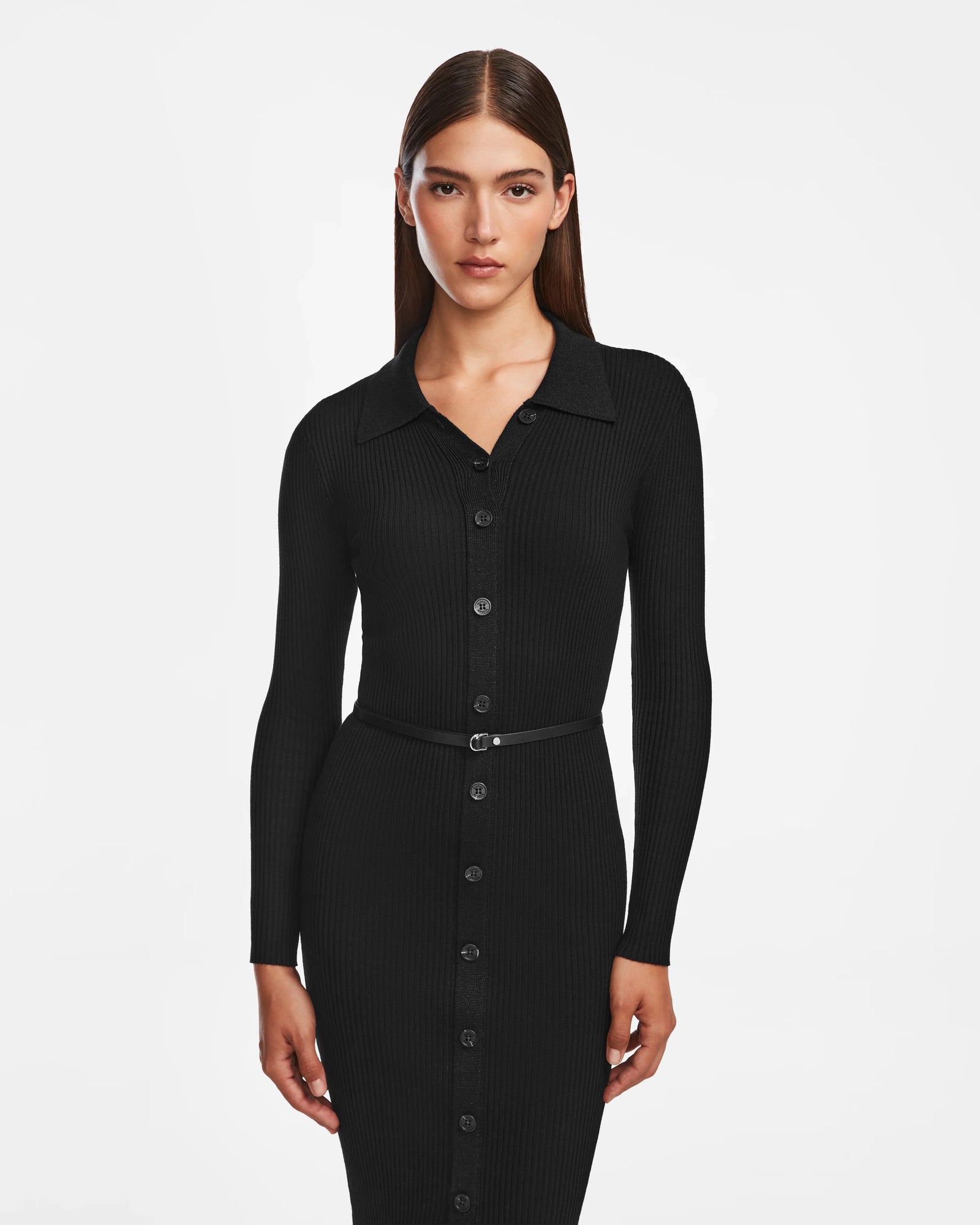 A woman with straight brown hair models the Rudsak CARLI - 8524573 Black, a fitted long-sleeve knit dress with front buttons and a narrow vegan leather belt, standing against a plain white background.