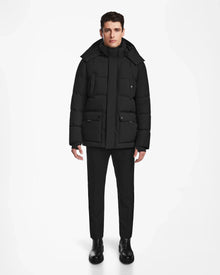 A person stands facing forward against a plain white background, wearing the Rudsak CARLOS - M124509 Black quilted parka coat with a hood, paired with black pants and black boots.