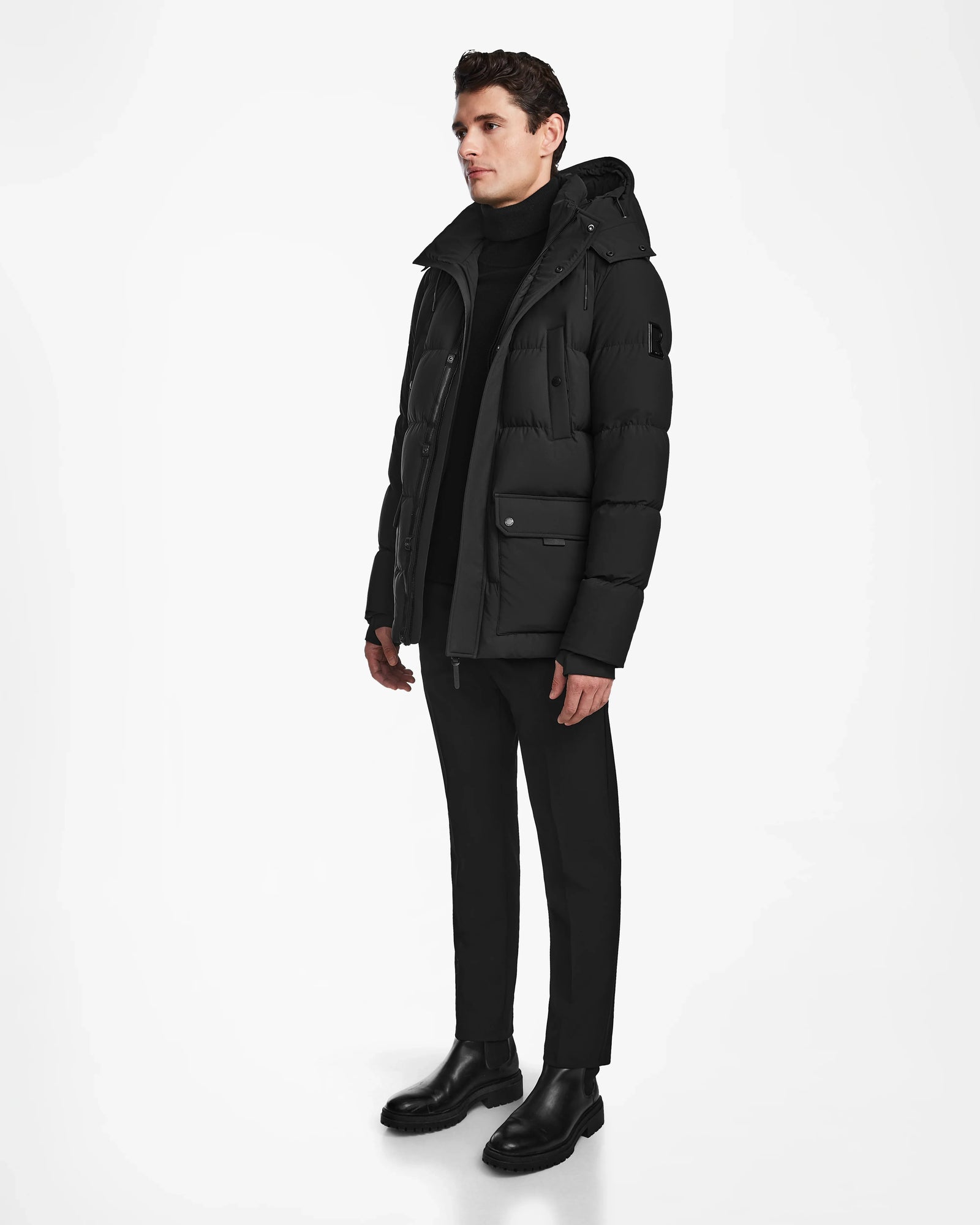 A man models the Rudsak CARLOS - M124509 Black parka against a white background, styled with a black turtleneck, trousers, and boots. He stands slightly turned to the side with his hands at his sides, highlighting the coats sleek winter design.