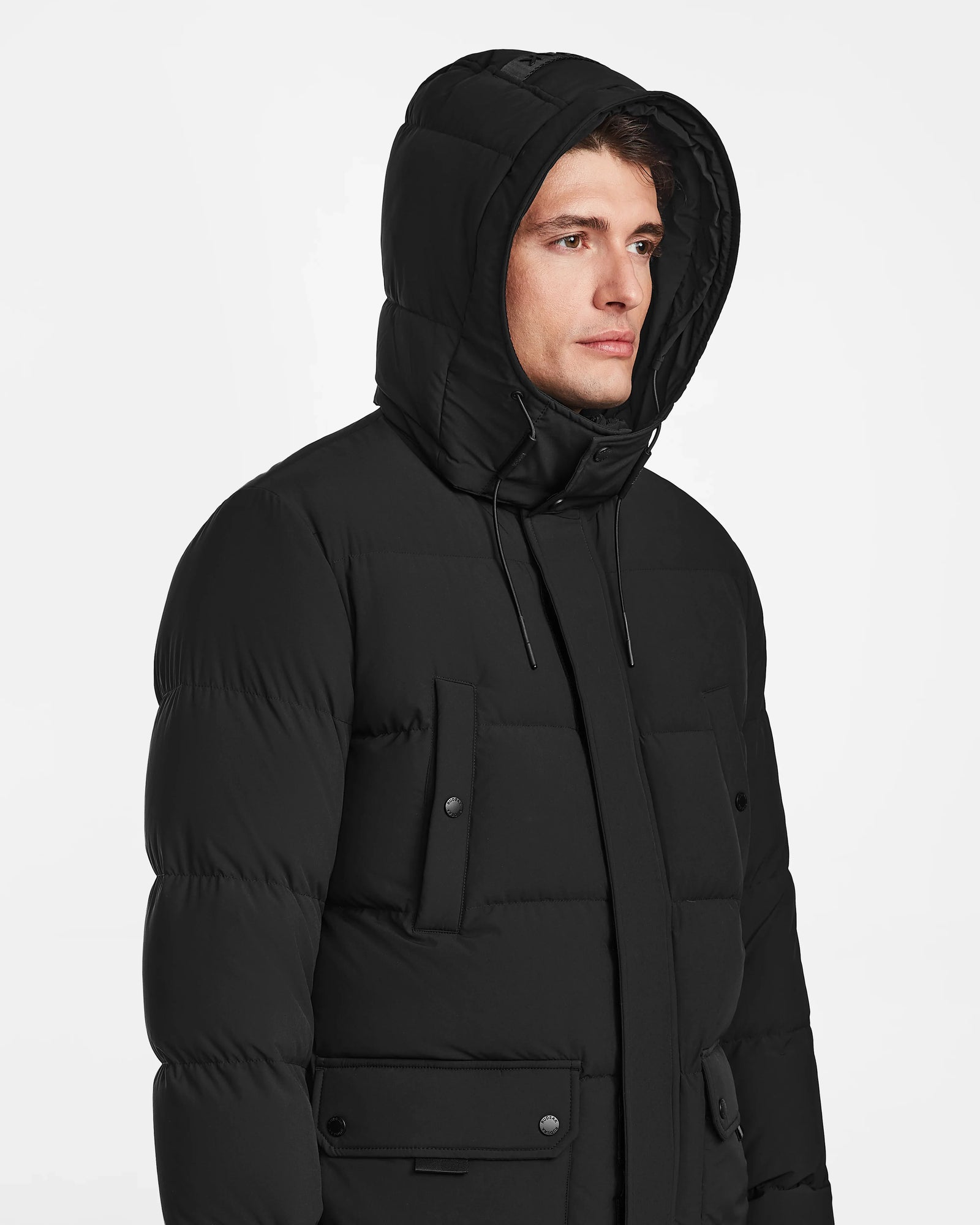 A man models the Rudsak CARLOS - M124509 Black quilted parka with a hood, large pockets, and button details, posing against a plain white background.