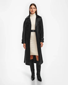 A woman stands facing forward in a black Rudsak CASSIE L trench coat (8124531) over a light beige turtleneck dress with a black belt, paired with knee-high black boots. The background is plain white.