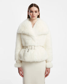 A woman faces forward wearing the Rudsak CHLOE - L124564 Iceland women’s faux fur jacket with wide lapels, a belted waist, and matching fur cuffs, styled with a white skirt against a plain white background.