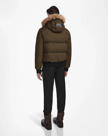 A person faces away, wearing the Rudsak DALLAS - M122501 Army weather-resistant puffer with a fur-lined hood, black pants, and brown boots against a plain white background.