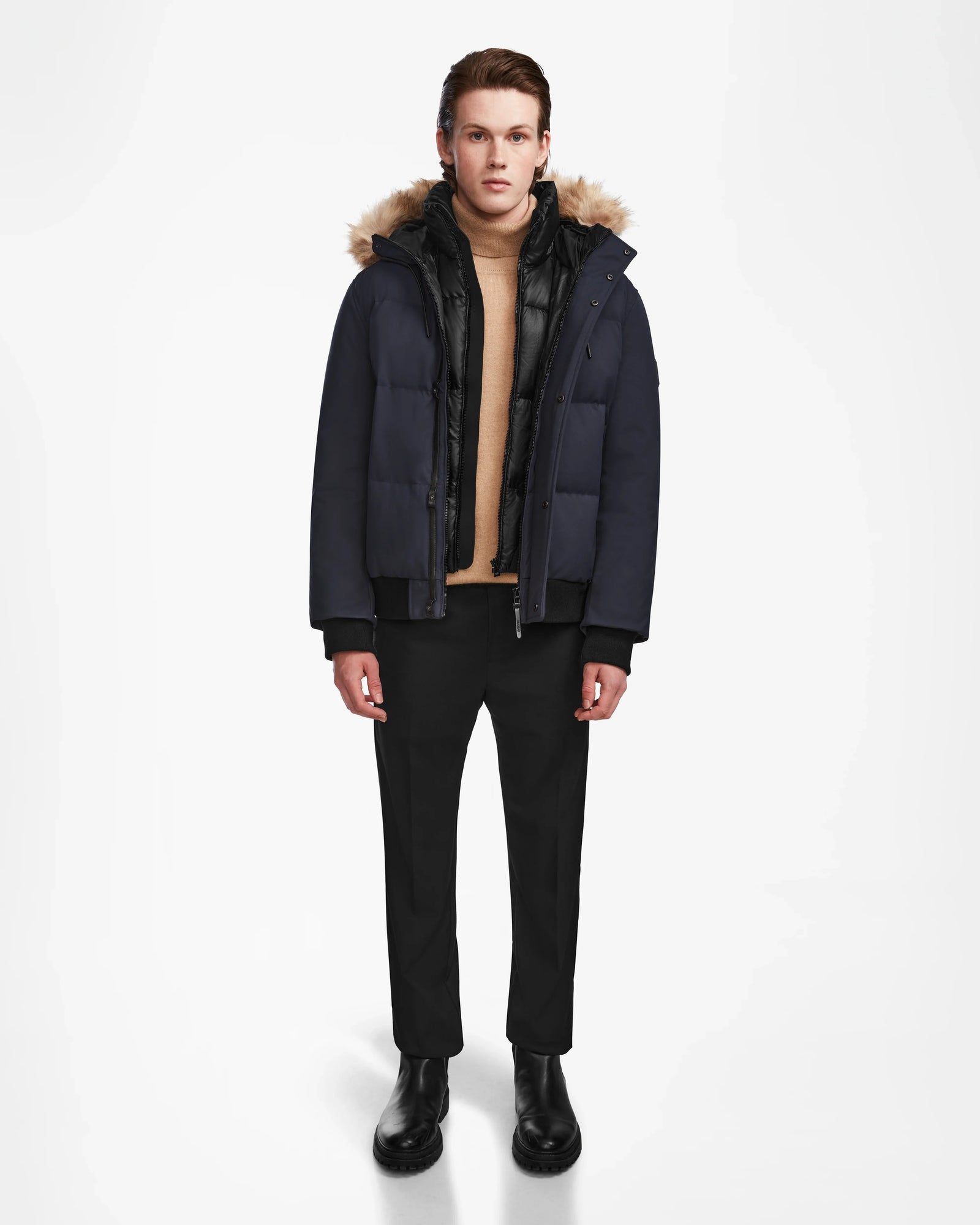 A person stands against a white background wearing the Rudsak DALLAS - M122501 Navy men's quilted down bomber with a fur-lined hood, paired with a beige turtleneck, black pants, and boots. The weather-resistant puffer offers warmth and style for winter.