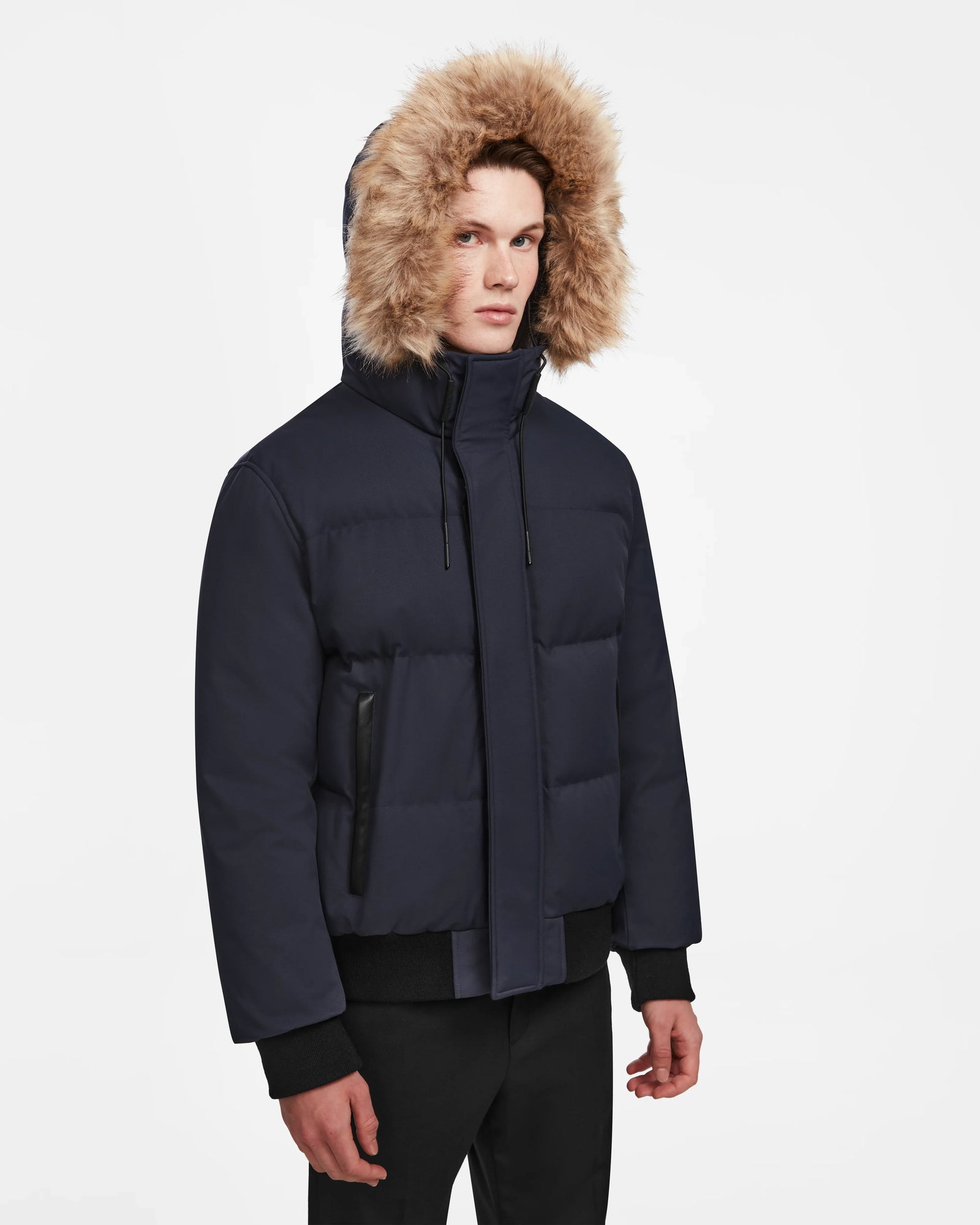 A young man in the Rudsak DALLAS - M122501 Navy weather-resistant puffer jacket with a fur-lined hood and black pants stands against a plain white background, wearing a neutral expression.