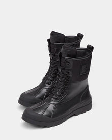The DAMIEN - M222500 Black by Rudsak are men's high-top winter boots in black, crafted from weather-treated leather with rugged soles, side zippers, stitched panels, and a raised R logo on the outer sides.