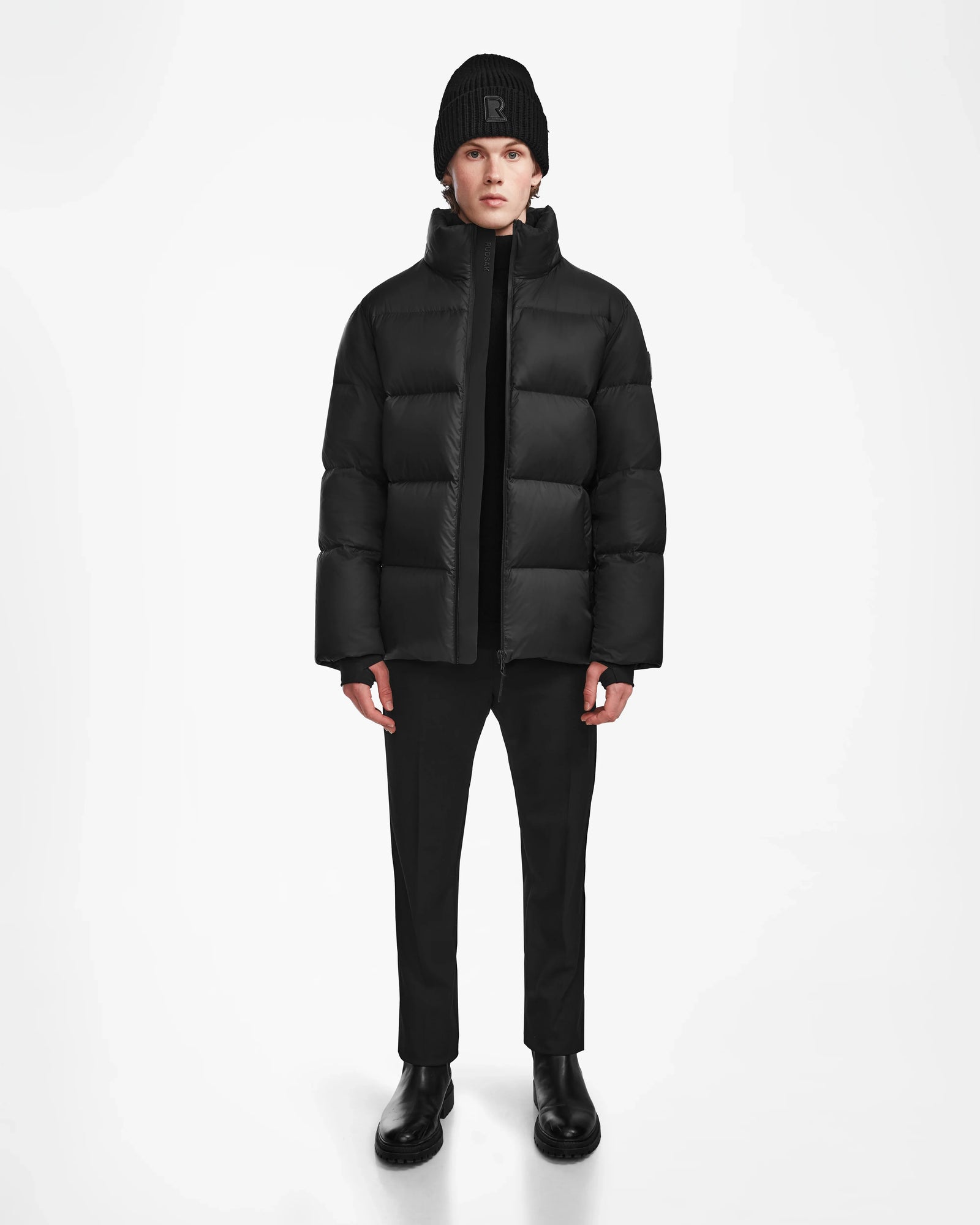 A person stands facing forward against a plain white background, wearing the Rudsak DANIEL M - M124510 Black weather-resistant puffer jacket with black pants, boots, and a beanie.