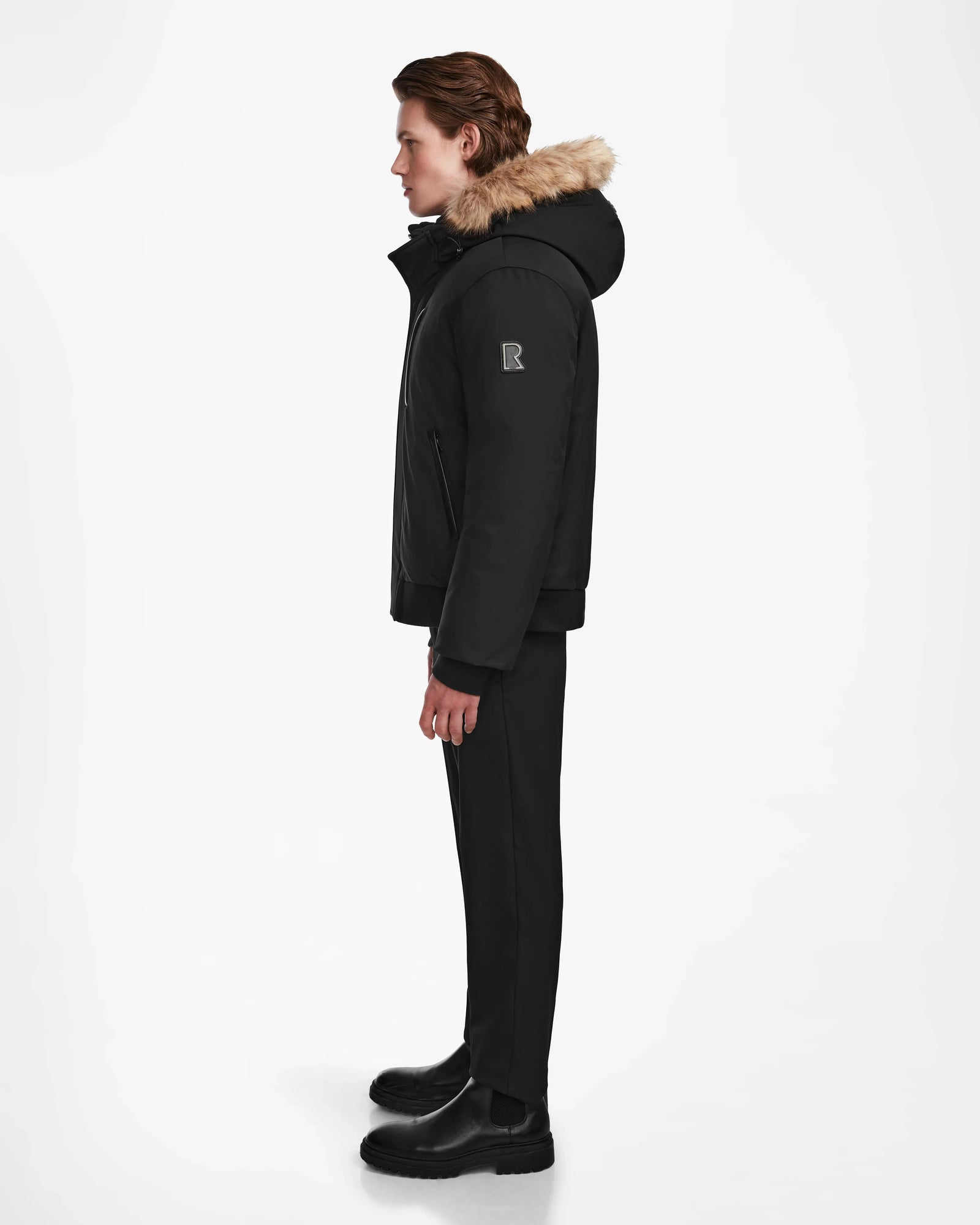 A person stands in profile facing left, wearing the Rudsak DEACON - M123513 Black/Natural down puffer jacket with fur trim, paired with black pants and boots against a plain white background.