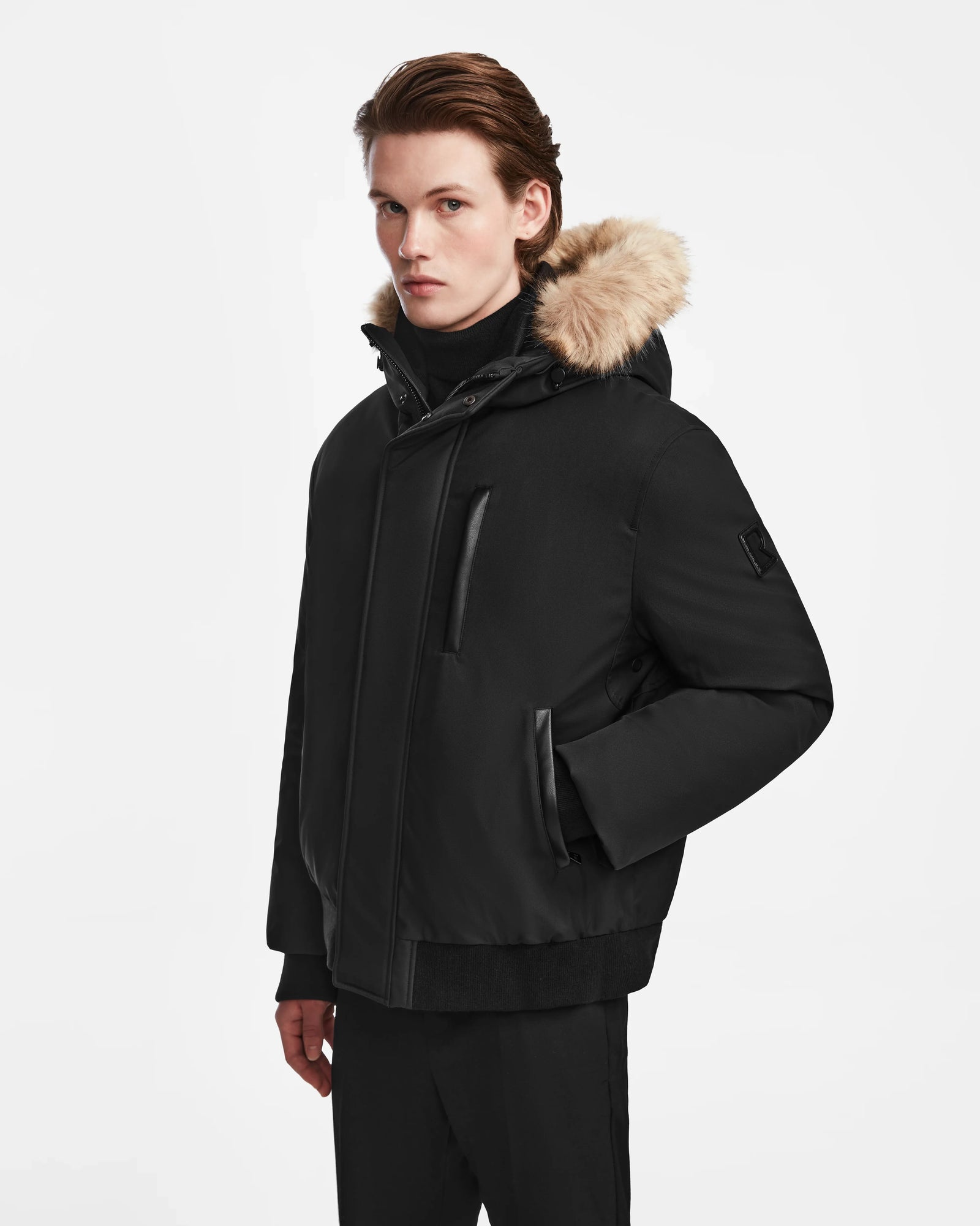 A young man with light skin and brown hair wears the Rudsak DEACON - M123513 Black/Natural down puffer with a fur-lined hood, standing against a plain white background and looking slightly to the side with a neutral expression.