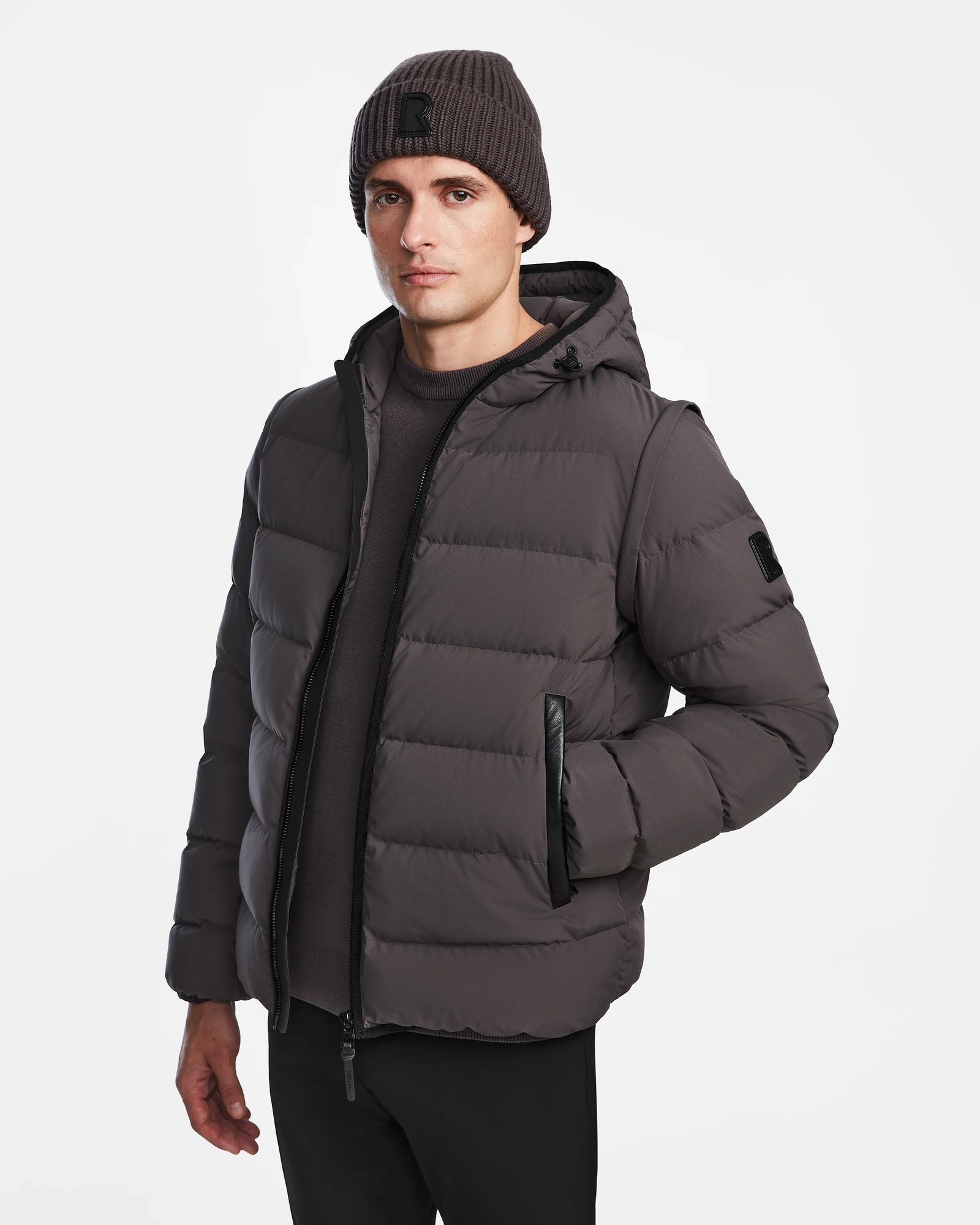 A person wears the Rudsak DEMIAN M-R - M123504 grey mens down puffer jacket made from sustainable recycled fabric, along with a gray beanie and black pants, standing against a plain white background.
