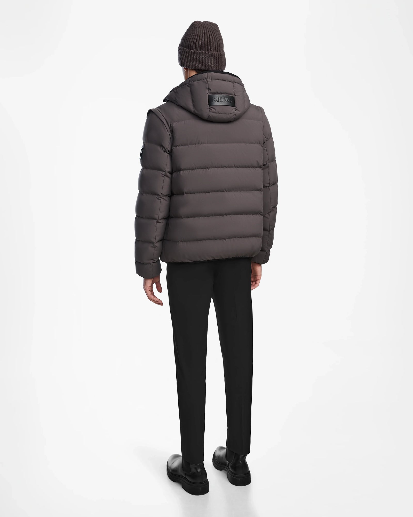 A person stands facing away, wearing the Rudsak DEMIAN M-R - M123504 Grey men’s down puffer jacket with a hood, paired with black pants, black shoes, and a brown knit beanie—all set against a plain white background.