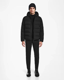 A person stands facing forward in the Rudsak DEMIAN M - M123507 Black hooded down puffer, made from sustainable recycled fabric, paired with black pants and boots, against a plain white background.