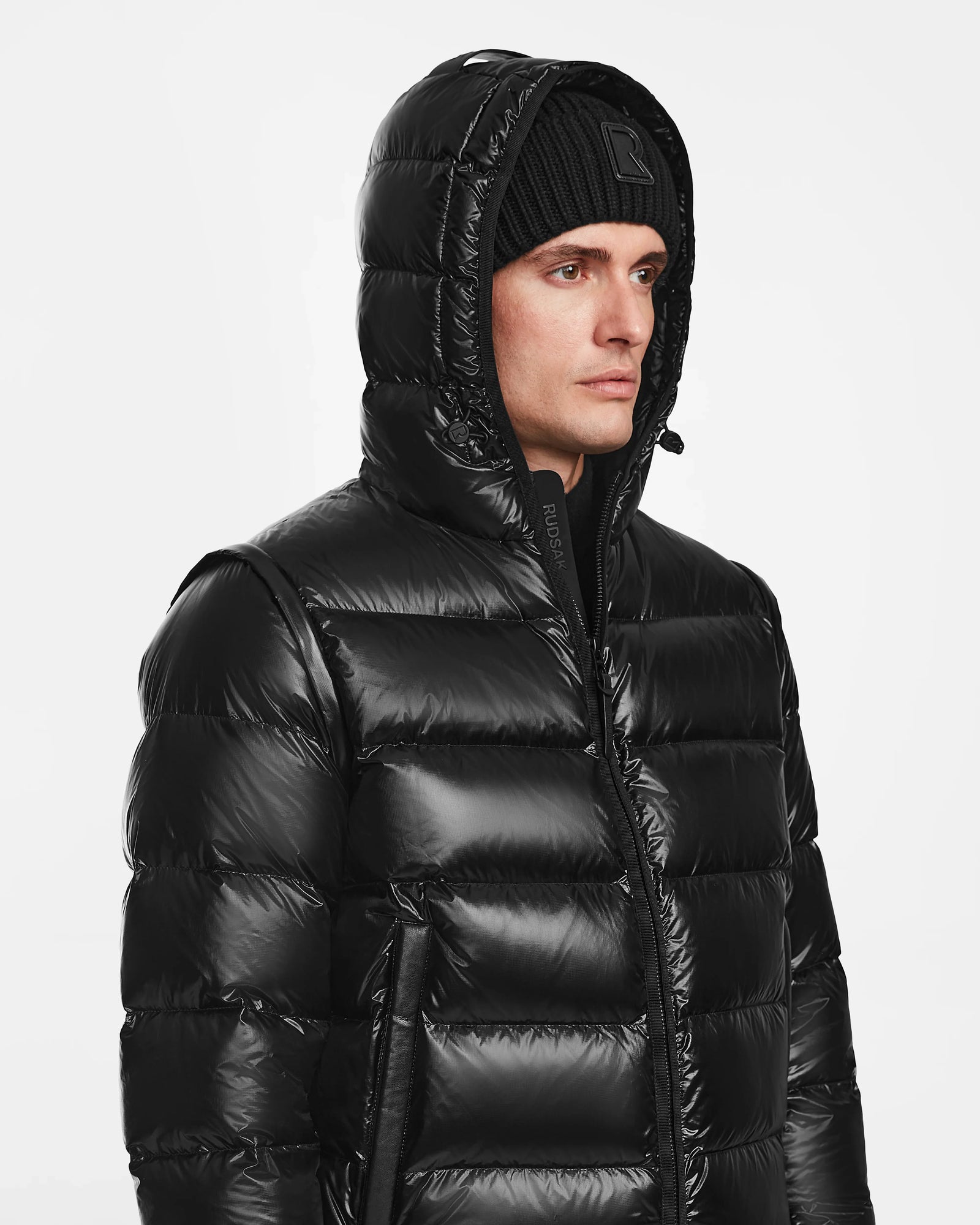 A man in the Rudsak DEMIAN S-R - M123505 Black down puffer jacket with hood up and a black knit beanie, glancing to the side against a plain white background.