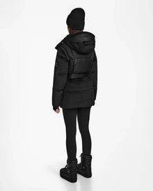 A person stands facing away, wearing the Rudsak DONIA - L124563 Black womens down puffer with leggings, platform boots, and a black beanie against a plain white background.