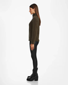 A woman with straight brown hair stands in profile, facing left, wearing the Rudsak EDITH - 8524617 Army vegan leather trim sweater with black leggings and chunky boots against a plain white background.
