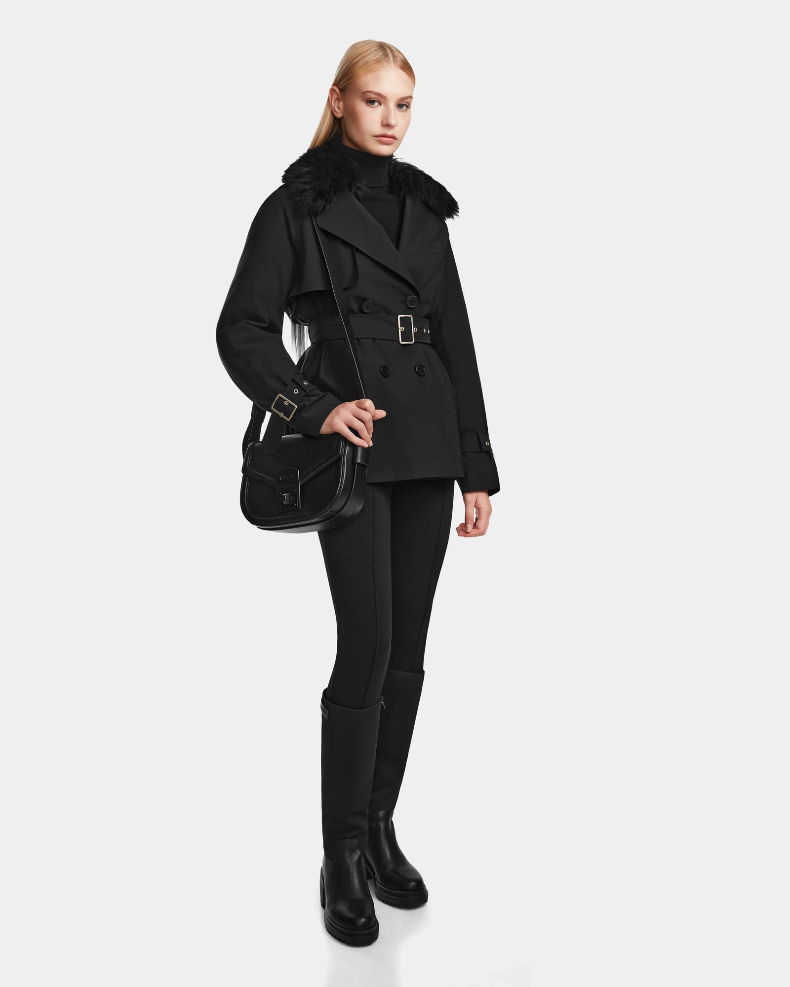 A woman stands against a plain white background wearing the Rudsak ELENOR - 8124707 Black trench coat with a shearling collar, paired with black pants, knee-high black boots, and a black crossbody bag.