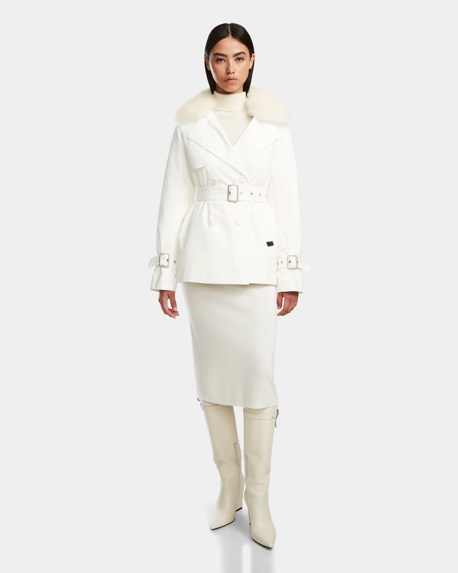 A woman wears the Rudsak ELENOR - 8124707 Iceland: a white down-filled trench with cozy shearling collar, paired with matching white boots.