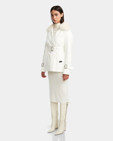 A woman models the Rudsak ELENOR - 8124707 Iceland white belted trench coat with shearling collar, paired with a cream turtleneck, matching midi skirt, and knee-high white boots against a light gray background.
