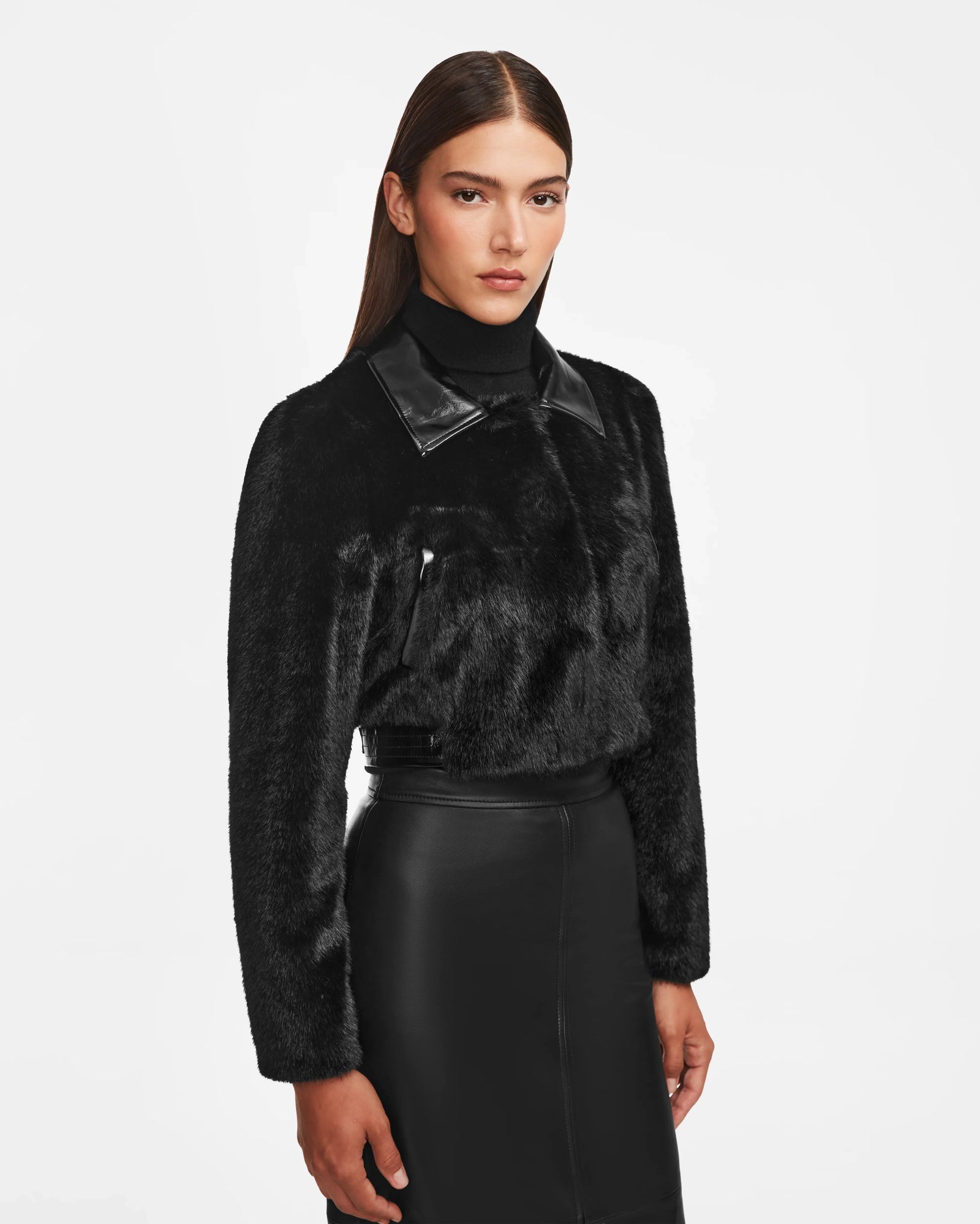 A woman models the Rudsak EVELYN - 8124791 Black faux fur jacket with a patent collar and black faux leather skirt, standing confidently against a white background in this striking, cropped fitted look.