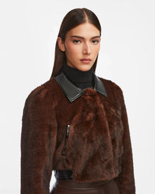 A woman with straight, dark hair wears the Rudsak EVELYN - 8124791 Espresso cropped faux fur jacket with a black leather collar over a black turtleneck, standing against a plain white background.