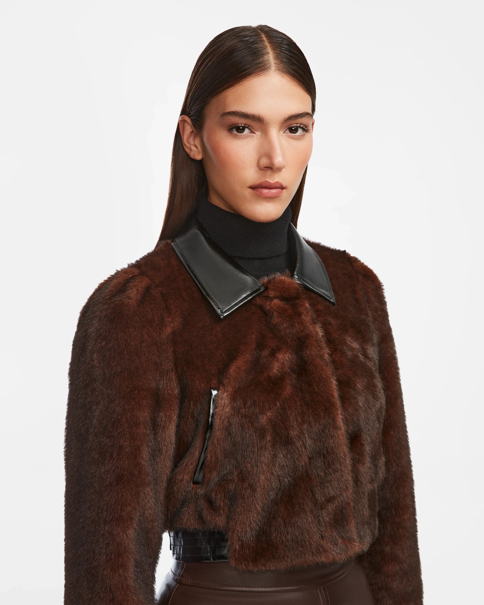 A woman with straight, dark hair wears the Rudsak EVELYN - 8124791 Espresso cropped faux fur jacket with a black leather collar over a black turtleneck, standing against a plain white background.