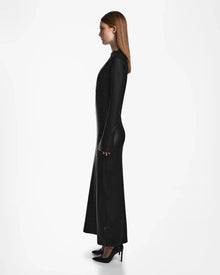 A woman with straight brown hair stands in profile against a white background, wearing the FAIZA - 8524579 Black maxi-length dress by Rudsak, styled with long sleeves and paired with black high-heeled shoes.